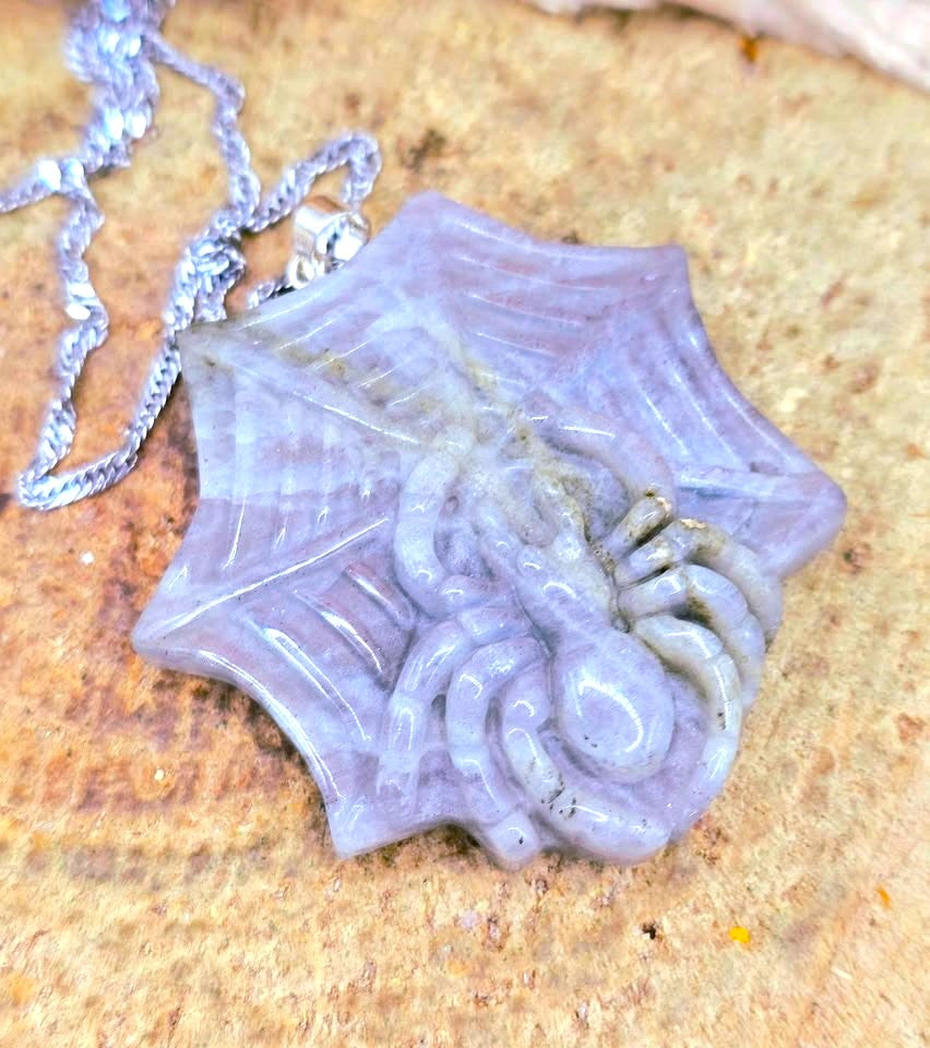 A spiderweb pendant necklace made of polished lavender-gray stone, hanging from a silver chain on a textured tan surface.