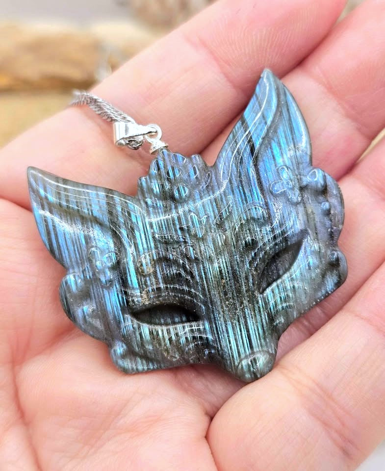 A hand holding a carved labradorite pendant shaped like a stylized fox or bird head with a silver chain.