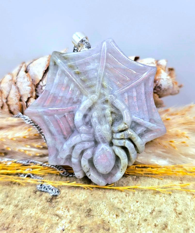A spider pendant necklace with a web design, made from a translucent pale lavender-gray stone, shown on a chain with decorative natural elements in the background.