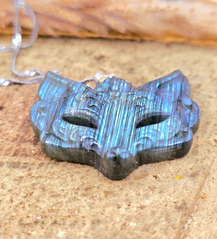 A carved mask pendant with a metallic blue and black iridescent finish, resting on a textured stone surface with a silver chain partially visible.