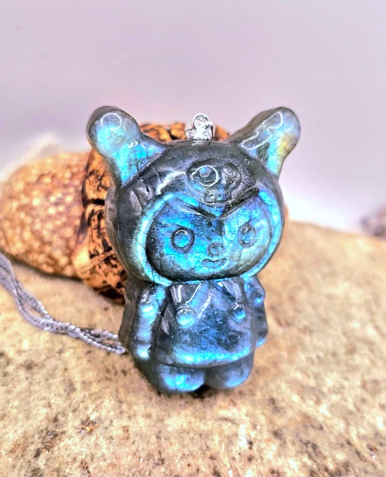 Natural labradorite pendant featuring a Kuromi‑inspired carving with smile and skull detail with silver chain displays on wood slice at side view-My Aura Gemstones