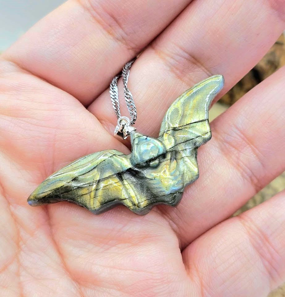 A close-up of a handmade pendant necklace featuring a carved bat-shaped piece made from layered green and blue stone with black veins, attached to a silver chain, resting on a textured tan stone surface.