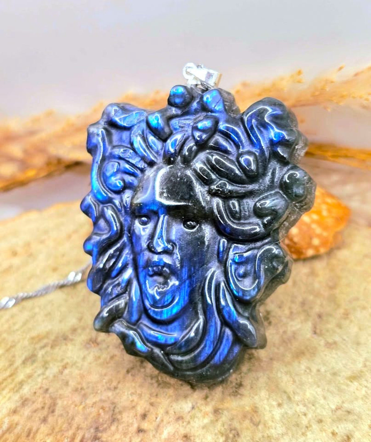 A labradorite pendant necklace featuring a sculpted face with swirling hair, displaying a vibrant blue and black iridescent play-of-color, resting on a textured stone surface.