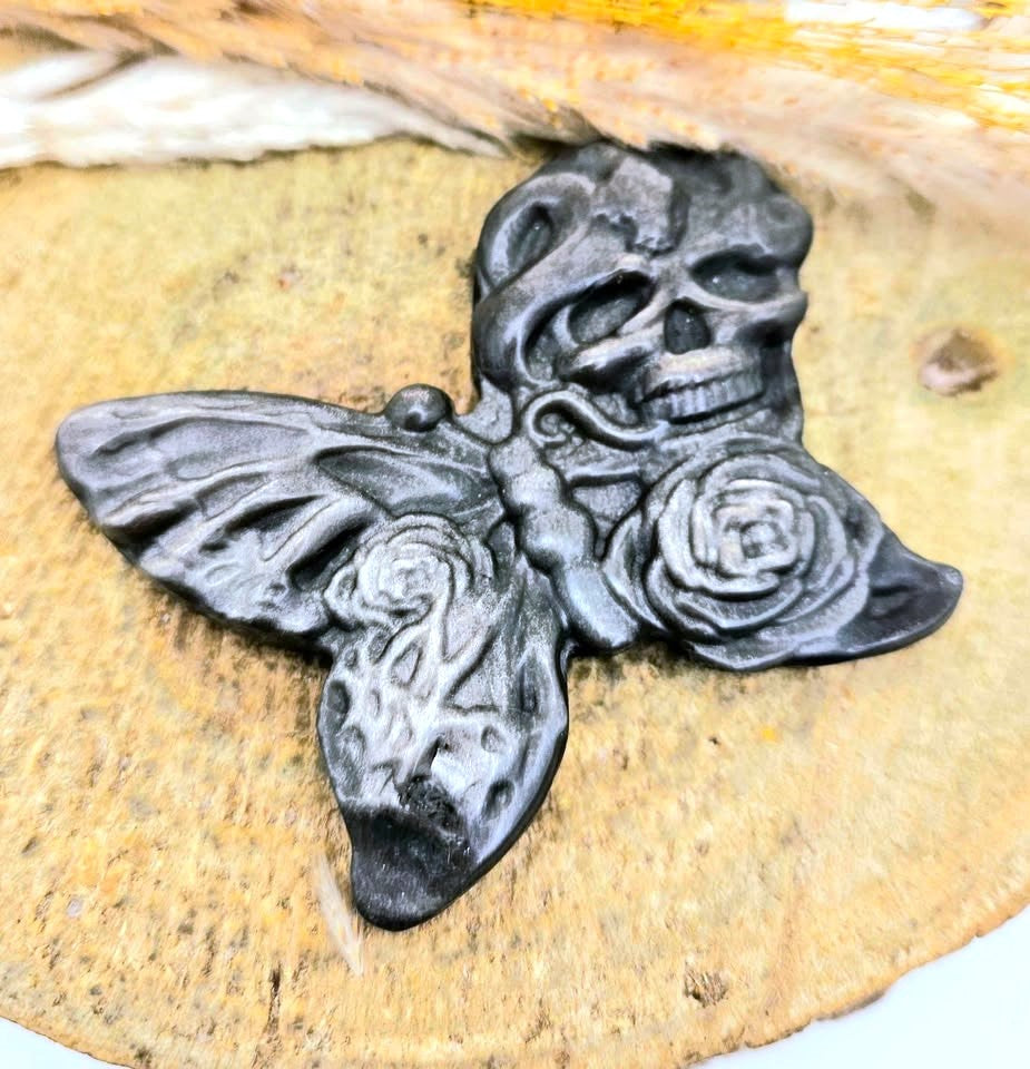 A black gothic-style butterfly silver obsidian featuring a skull on one wing and roses on the other, displayed on a textured tan stone surface.