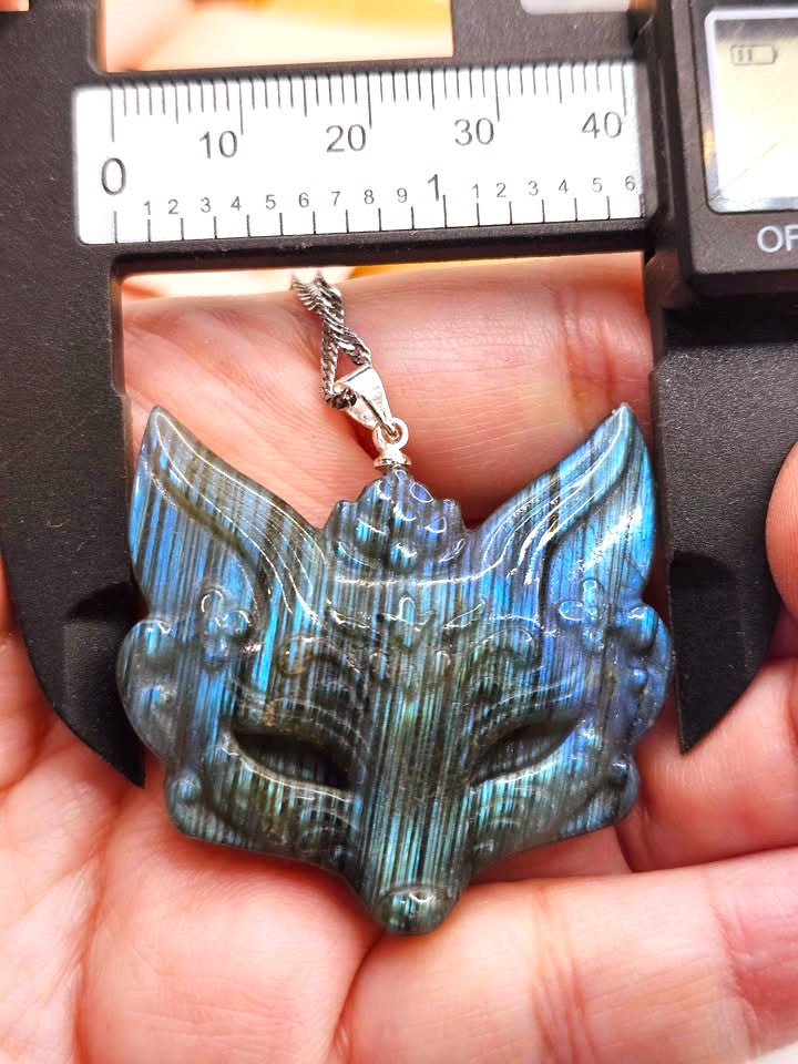 A hand holds a carved pendant shaped like a fox head, made from iridescent blue-green stone with a silver clasp, while a caliper is positioned above it for size reference.