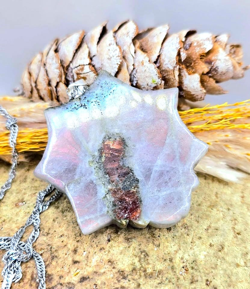 A star-shaped pendant necklace made of iridescent silver-toned metal with a central copper and dark oxidized inlay, displayed on a textured stone surface with a pinecone and dried grass in the background.