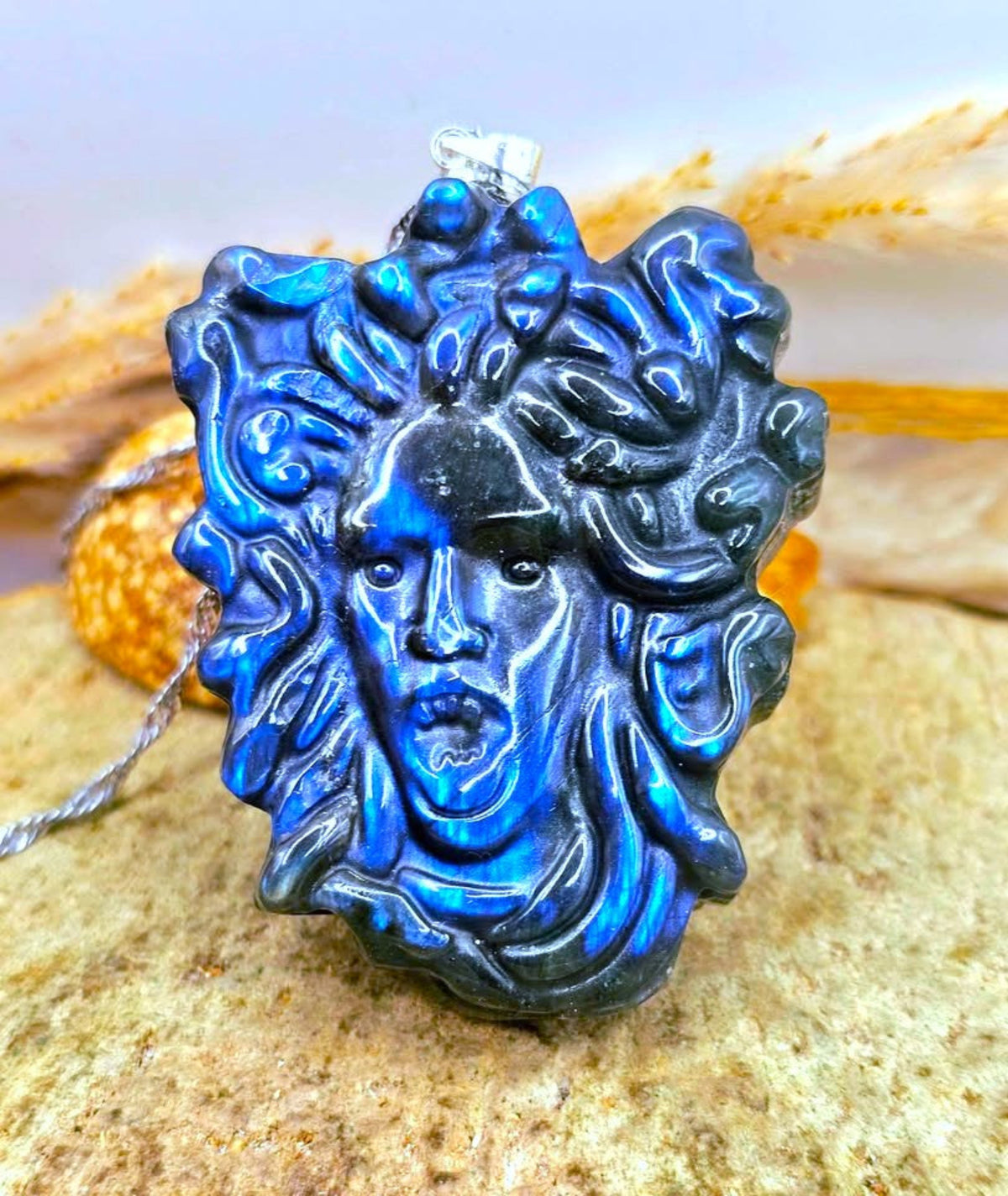 A blue and black polished stone pendant shaped like a Medusa head with flowing hair, resting on a textured beige rock with dried grass in the background.