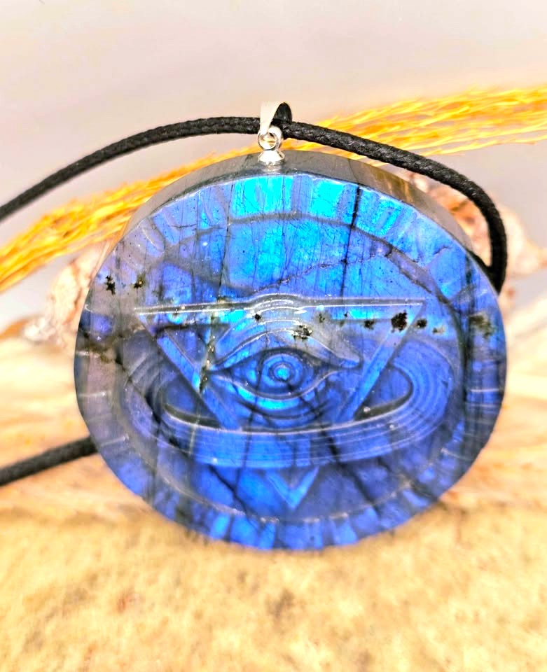 A round blue labradorite pendant featuring a carved eye and triangle design, suspended on a black cord, set against a textured beige background with gold-colored decorative elements.