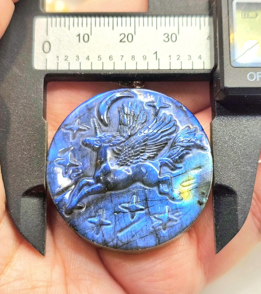 A round blue iridescent pendant or medallion with a carved winged horse and stars, held in a hand under a caliper to show size. The pendant has a deep blue lustrous finish with lighter blue and yellow-green shifts.