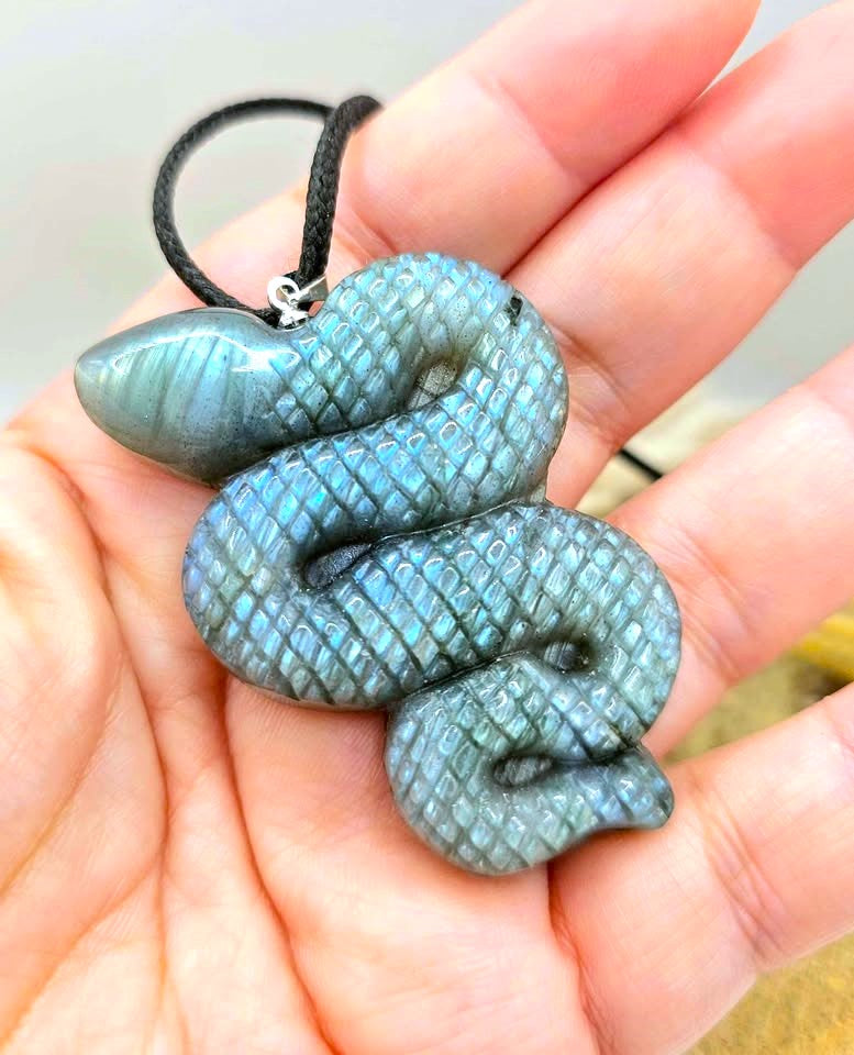 A coiled snake-shaped pendant made of iridescent labradorite stone, resting on a person's palm. The pendant has a black cord and a small silver clasp.