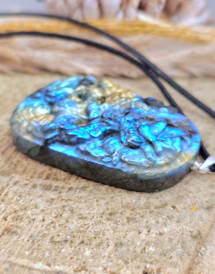 A carved labradorite pendant necklace with a detailed relief design, featuring vibrant blue and green iridescent flashes, resting on a textured stone surface with a black cord.
