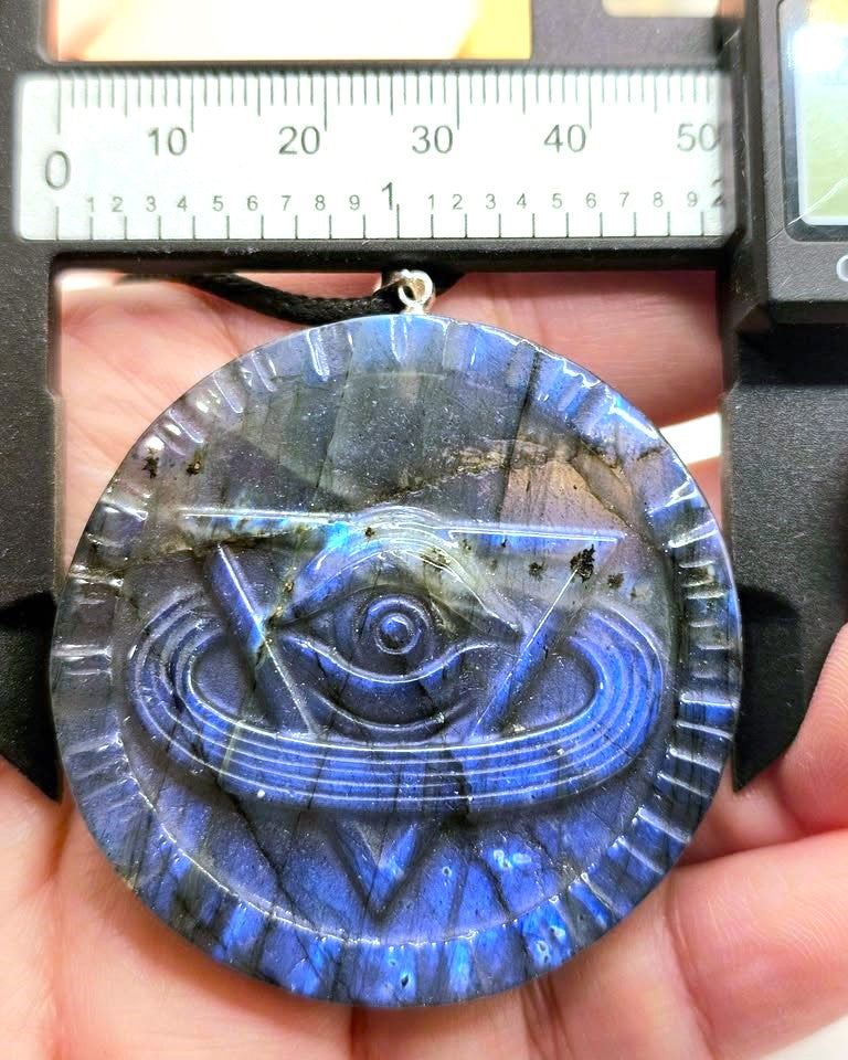 A round labradorite pendant with a carved eye design, held in a hand under a caliper to show its size. The pendant glows with blue and purple iridescence.
