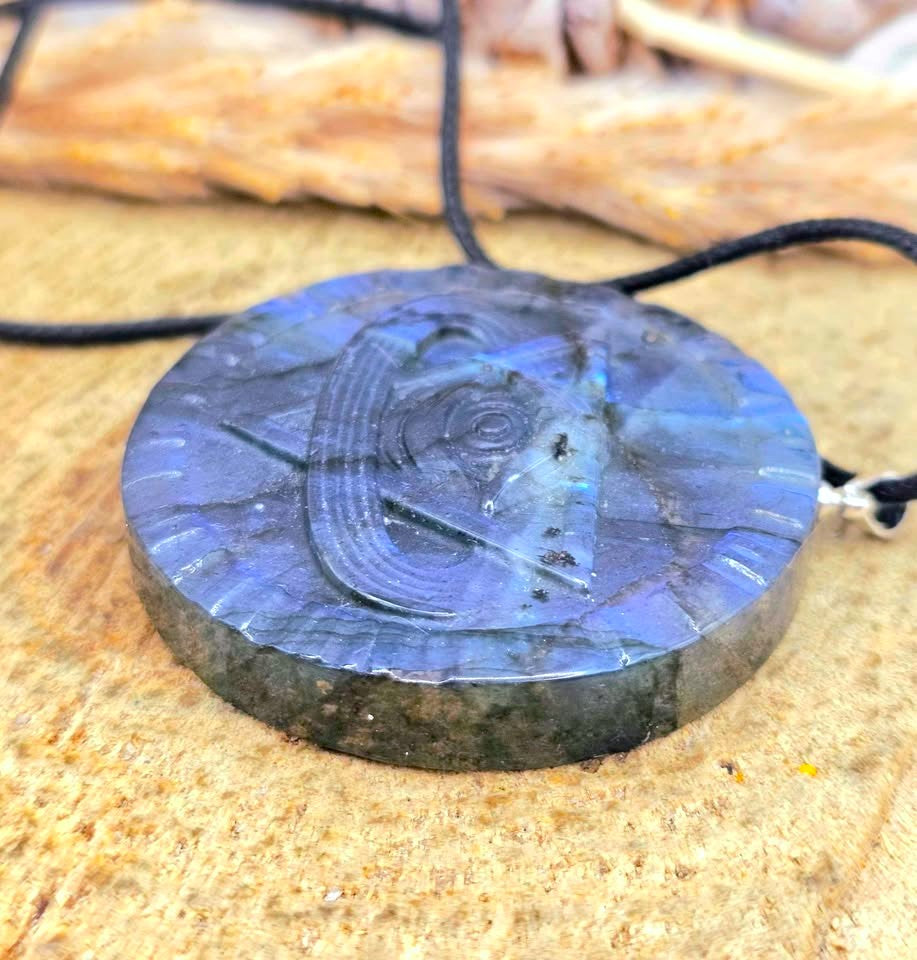 A round labradorite stone pendant necklace with a carved Polynesian-style wave design, resting on a textured tan surface with a black cord.