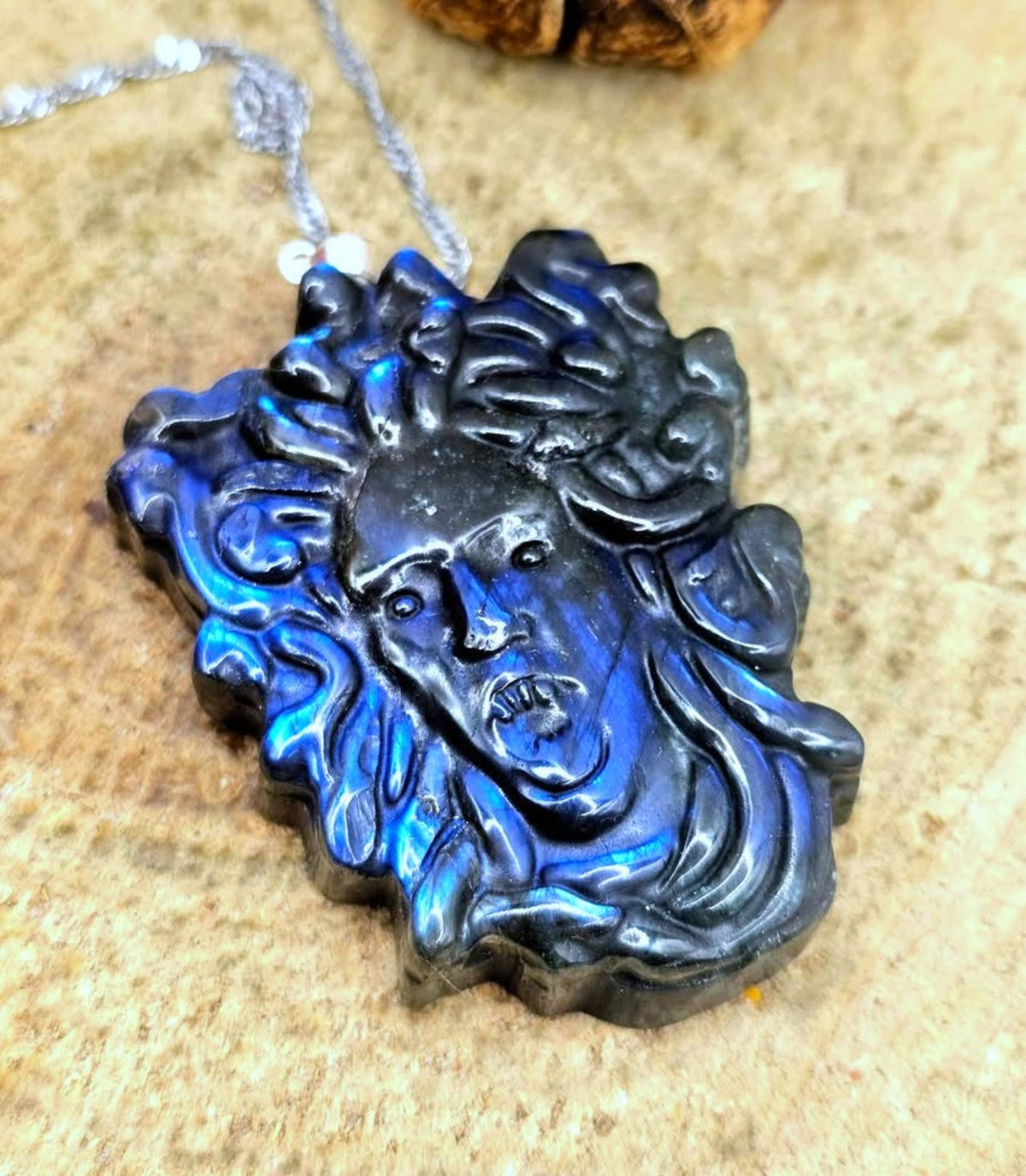 A carved black stone pendant with a blue iridescent sheen, depicting a Medusa-style face with flowing hair, hanging from a silver chain on a textured beige surface.