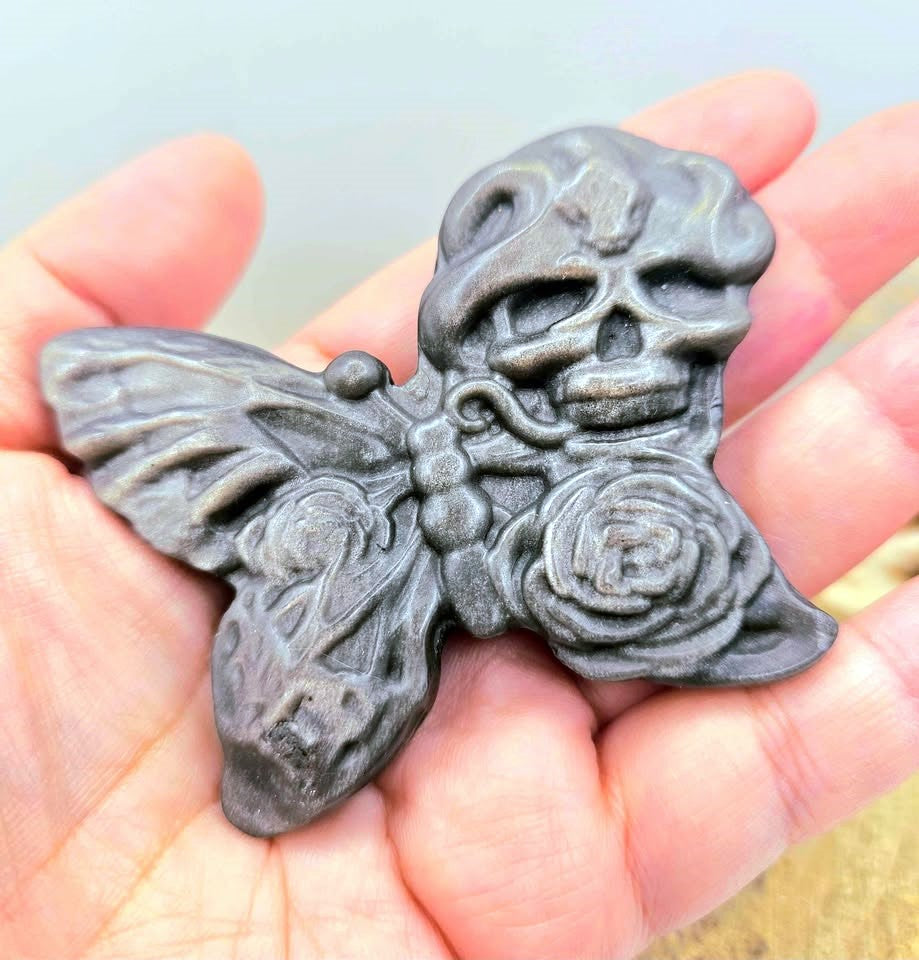 A dark gray gothic silver obsidian stone slice carved with butterfly with a skull on one wing and a rose on the other, resting on human hand.-My Aura Gemstones
