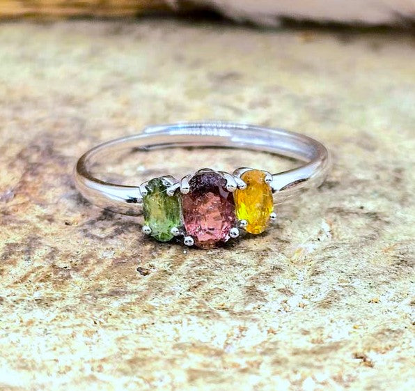 A silver three-stone ring featuring oval-cut green, pink, and yellow gemstones, displayed on a textured beige stone surface.