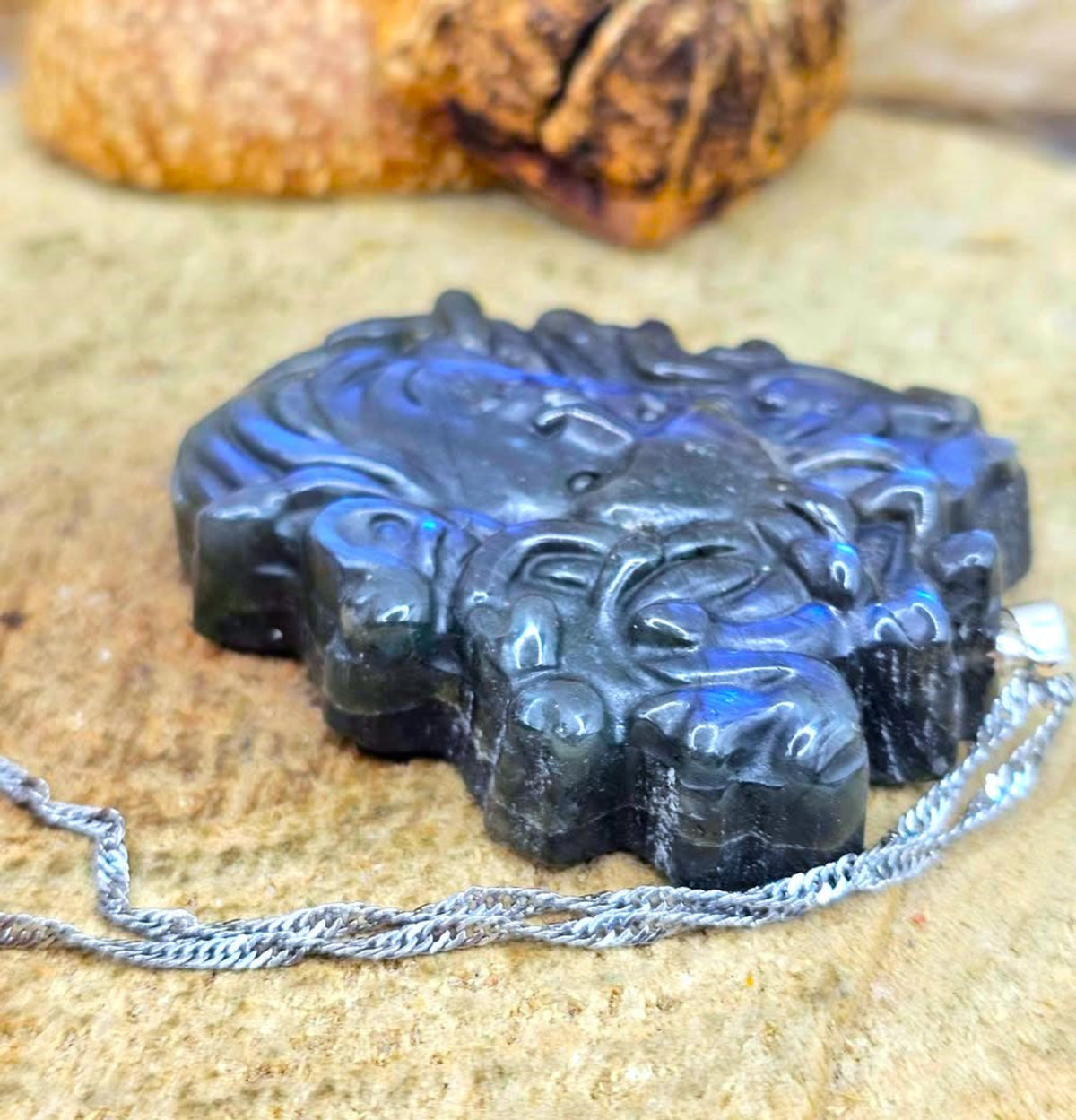 A carved black labradorite pendant with a silver chain, displayed on a textured stone surface with a blurred nut in the background.