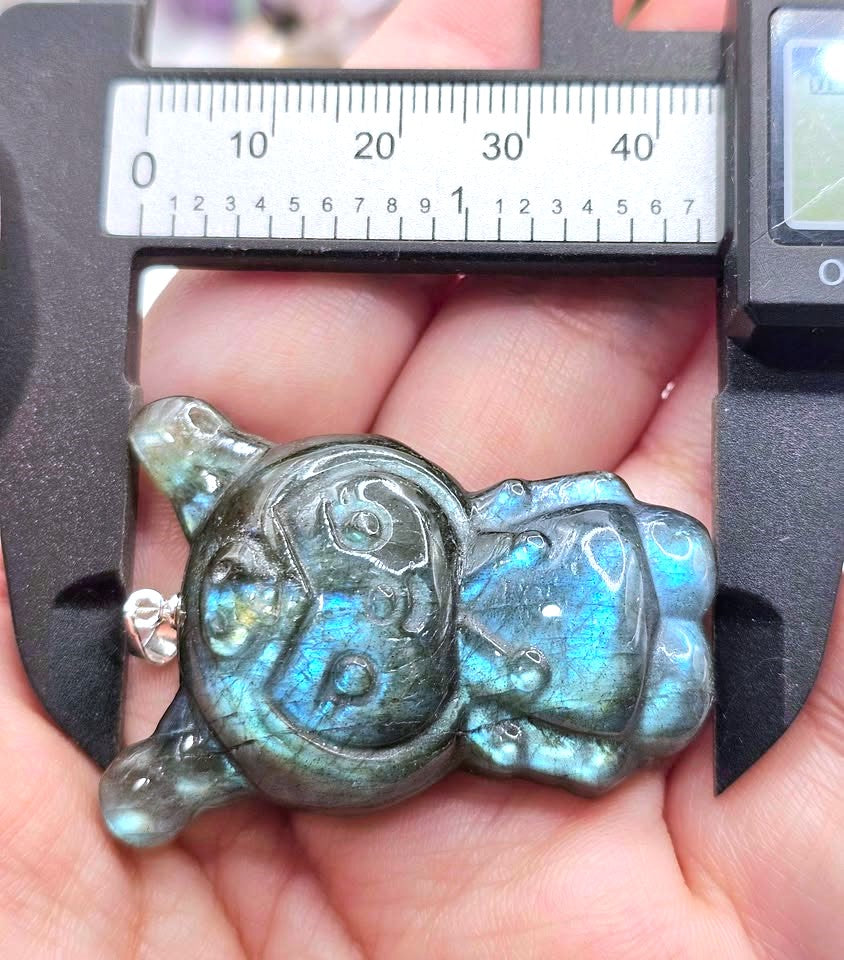 A carved labradorite pendant shaped like a smiling face, held in a person's hand under a caliper to show its dimensions. The pendant features a shimmering blue-green labradorite finish.