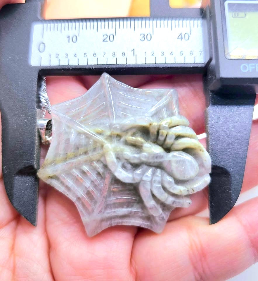 A small spider-shaped translucent resin charm held in a human hand under a digital caliper to show its size. The spider is detailed with green and brown accents on a clear, web-like base.