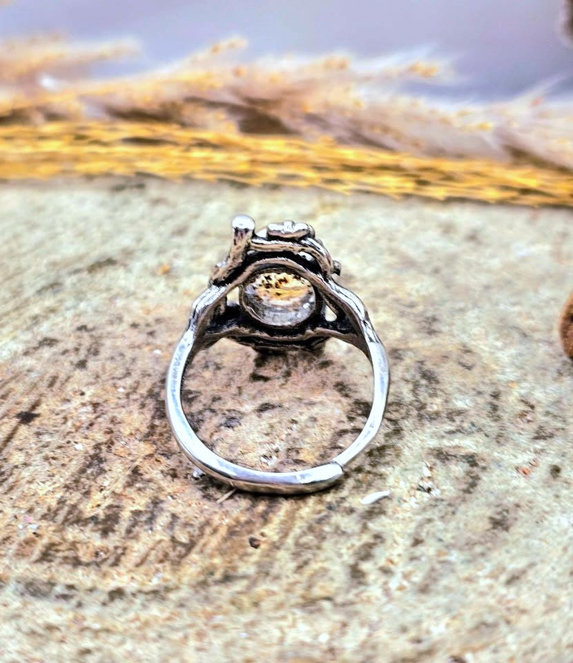 A silver-toned ring with an ornate openwork band and a central bezel-set clear gemstone, photographed resting on a textured stone surface with a blurred natural background.