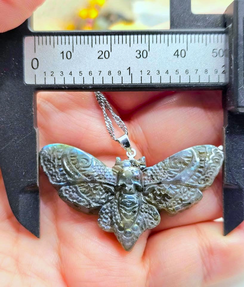 A silver moth pendant necklace held in a person's hand under a caliper to show its size. The pendant features intricate, textured wings and a silver chain.
