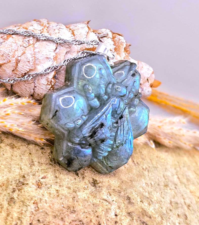 A close-up of a carved labradorite pendant necklace with a silver-toned chain, resting on a textured stone surface with dried grass and a pinecone in the background.