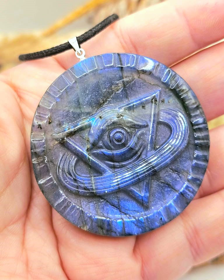 A round labradorite pendant necklace held in a hand. The pendant features a carved blue eye and triangle design with a silver bail and black cord.
