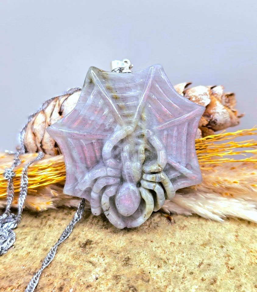 A spider-shaped pendant necklace with a web design, featuring a pale pink and gray stone body with a silver-toned chain, displayed on a textured surface with dried grass and pinecones.