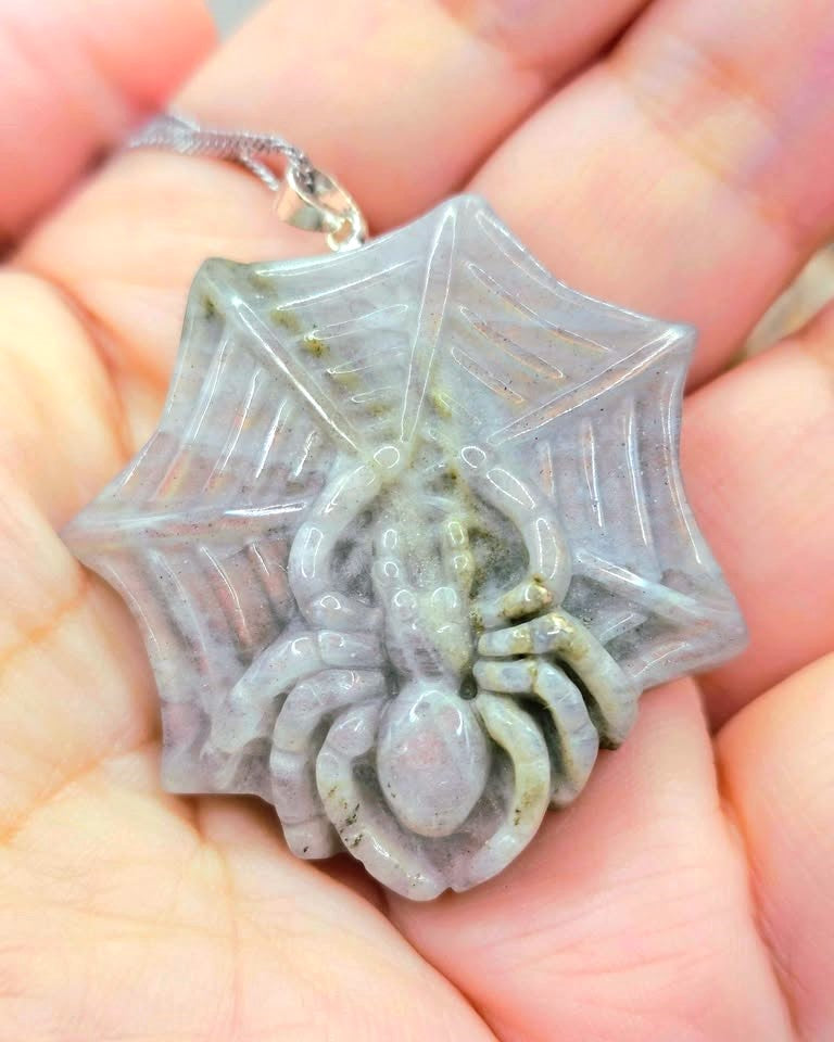 A carved spider pendant on a web-shaped background, held in a person's hand. The pendant appears to be made of a pale gray stone or resin with subtle greenish-brown veining, and a silver-toned chain is visible at the top.