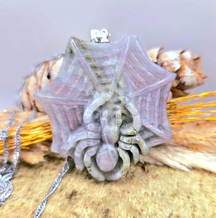 A spider-shaped pendant with a web design, made from a translucent pale lavender-gray resin with glitter, hanging from a silver chain. It is displayed on a textured surface with dried grass and pinecones.