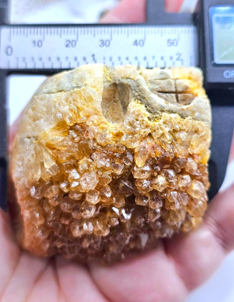 A hand holds a rough crystal specimen with clustered translucent brown and amber crystals on a tan matrix, with a caliper ruler visible behind it for scale.
