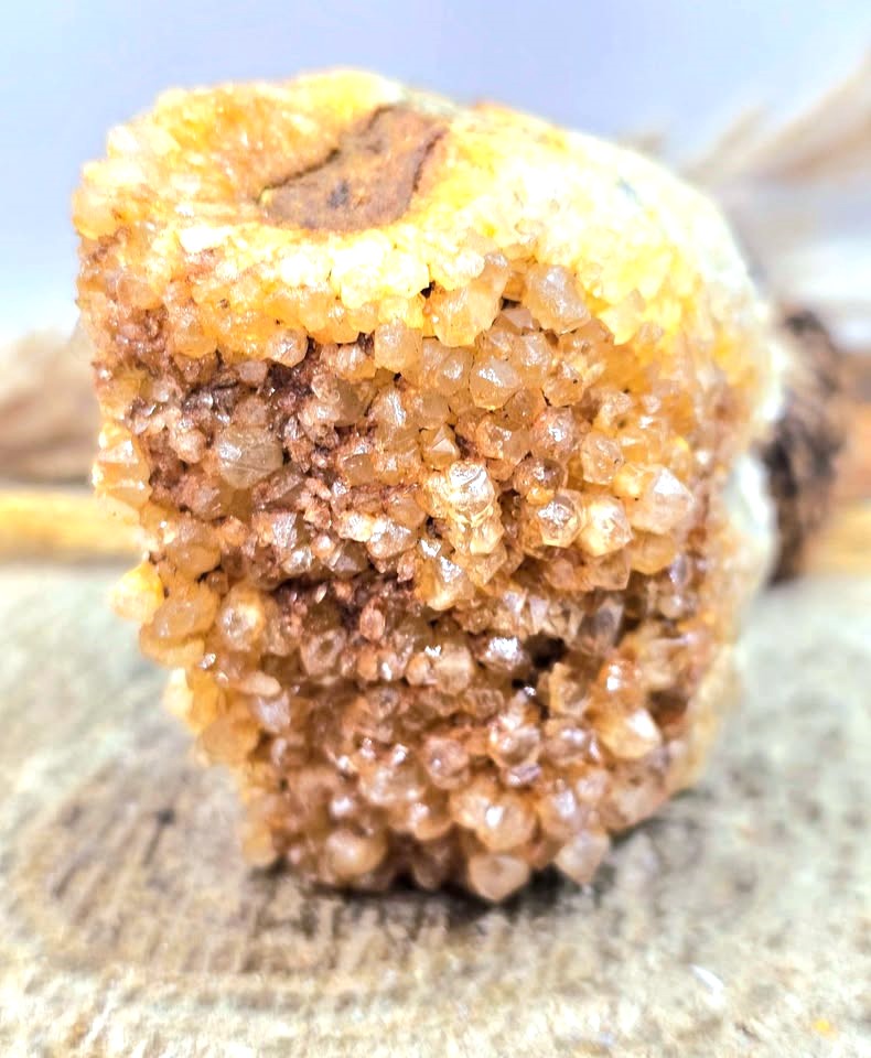 A close-up of a rough, textured crystal cluster with dense small faceted stones in warm amber, tan, and brown tones, photographed against a softly blurred neutral background.