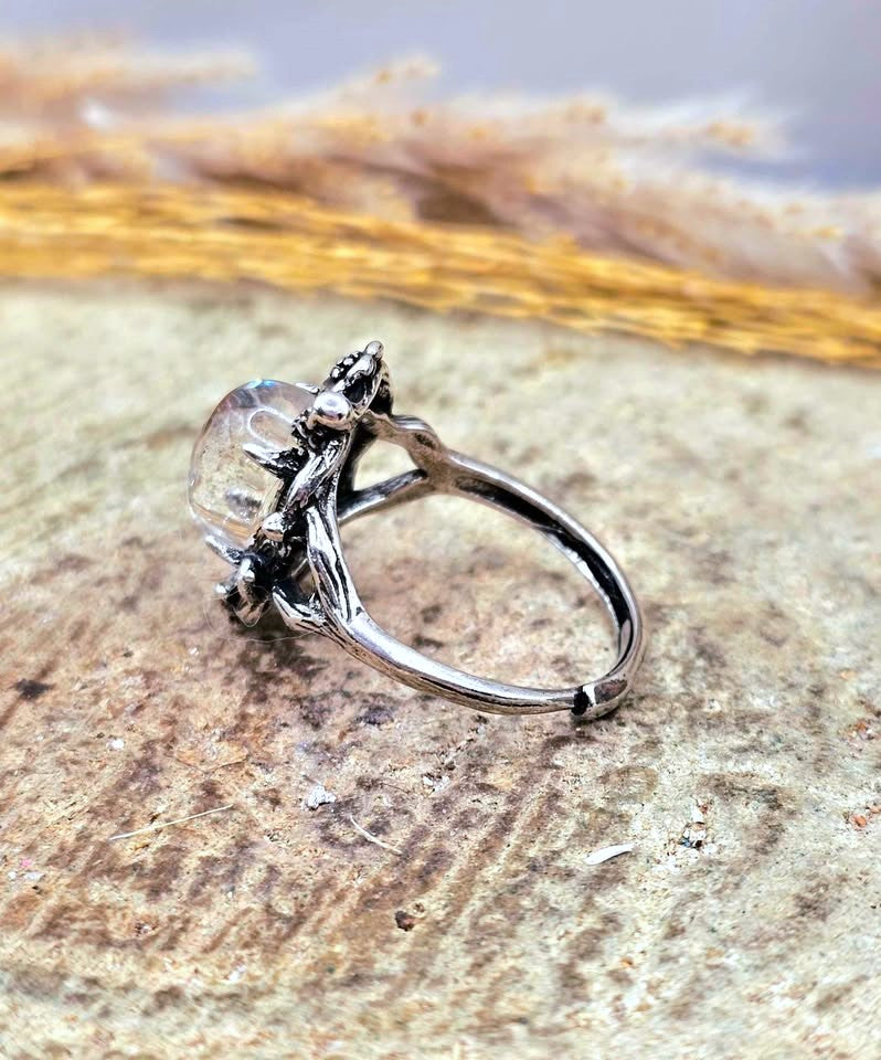 A silver ring with a clear faceted gemstone and organic, branch-like detailing, resting on a textured stone surface with dried grass in the background.