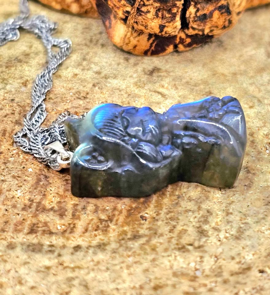 A blue iridescent pendant necklace shaped like a hooded figure, shown close up against a textured brown rock background with part of a silver chain visible in side view.-My Aura Gemstones
