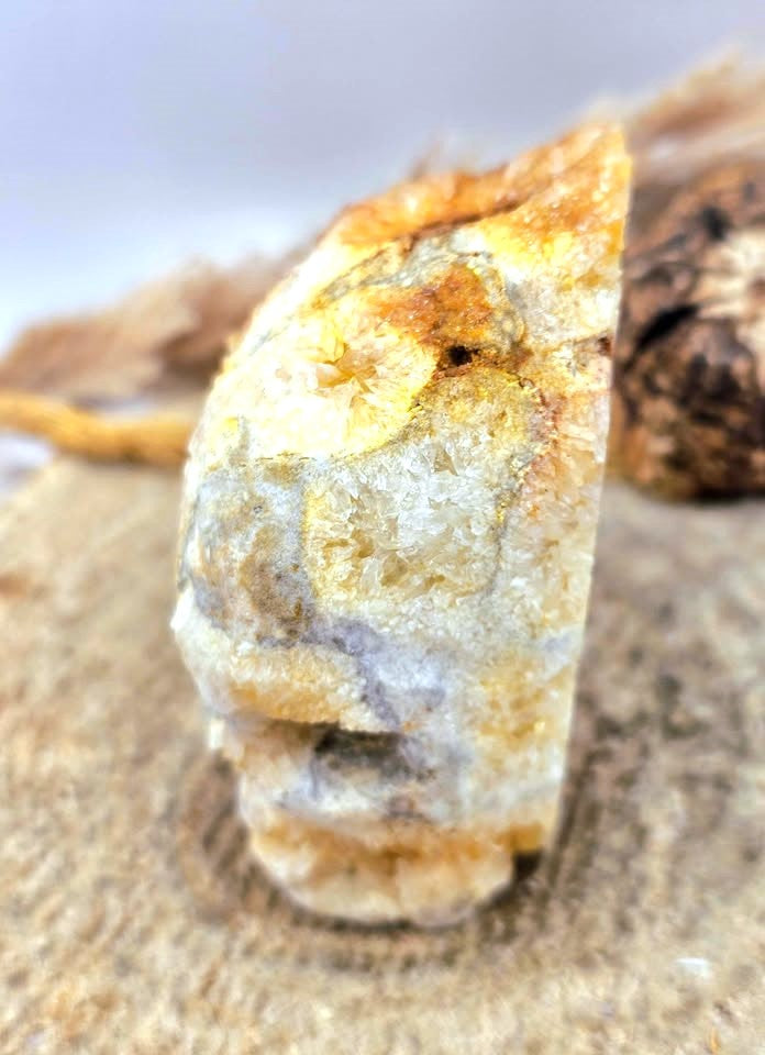 A close-up of a rough, natural crystal or mineral specimen with layered white, gray, and warm orange-brown tones, standing upright on a sandy surface with blurred natural background elements.
