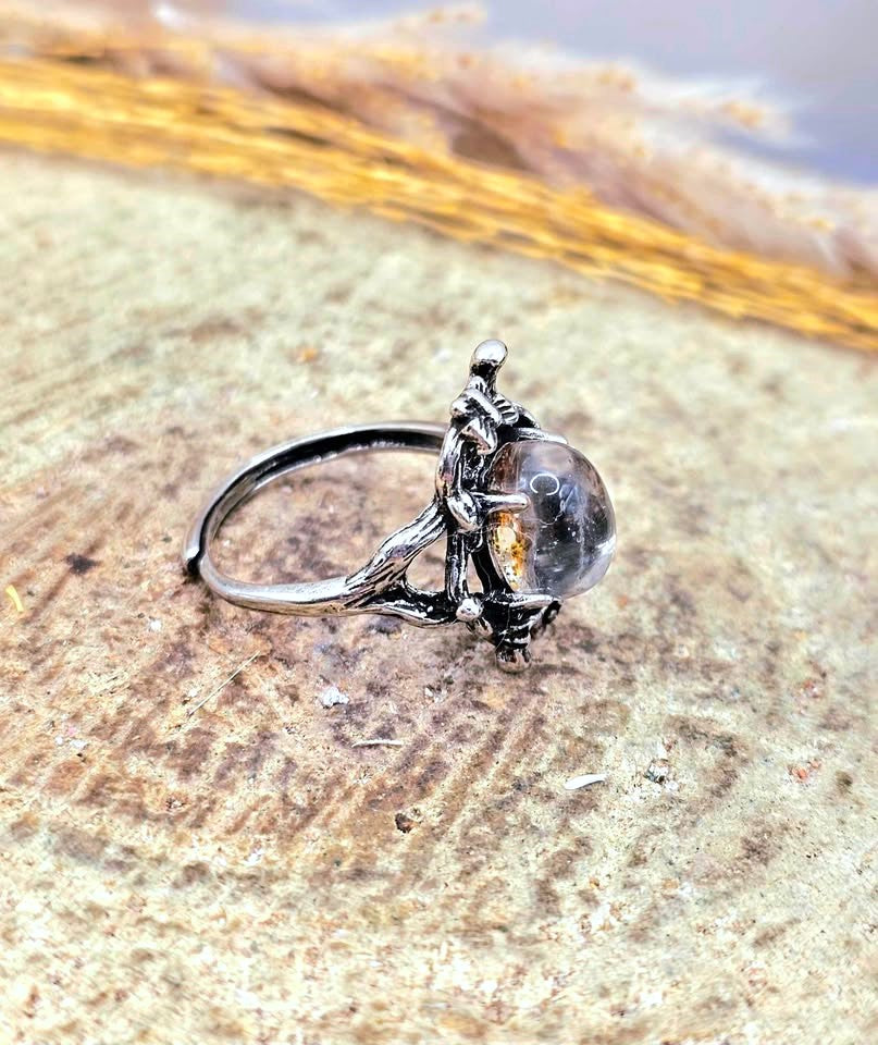 A silver-toned ring featuring a small sculpted figure holding a clear oval gemstone with a warm amber inlay, resting on a textured stone surface.