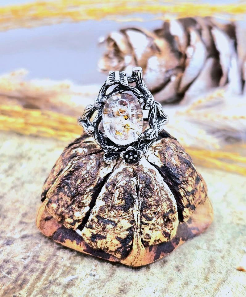 A gothic-style ring featuring a clear crystal cabochon with gold flecks, set in a detailed silver and black metal frame with a pumpkin-like base, resting on a textured surface.