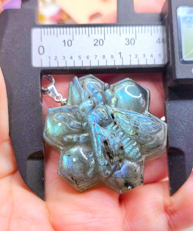 A close-up of a carved labradorite bee pendant held in a person's hand under a digital caliper to show its size. The pendant features a natural iridescent labradorite finish with blue, green, and gray flashes.