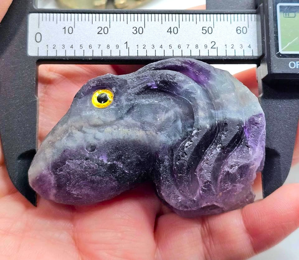 A raw, polished purple and gray stone carving shaped like a stylized animal head with a yellow eye, held in a hand under a digital caliper to show its size.