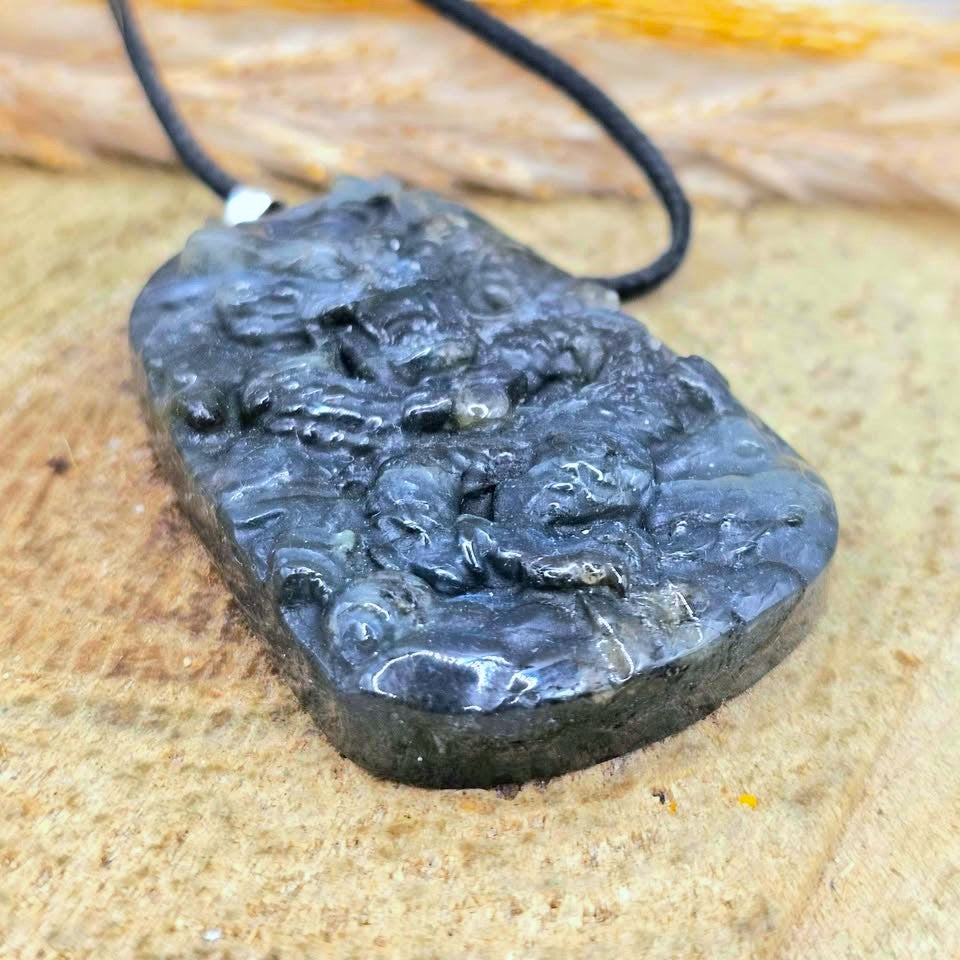 A dark gray carved stone pendant necklace with a black cord, resting on a textured tan surface.