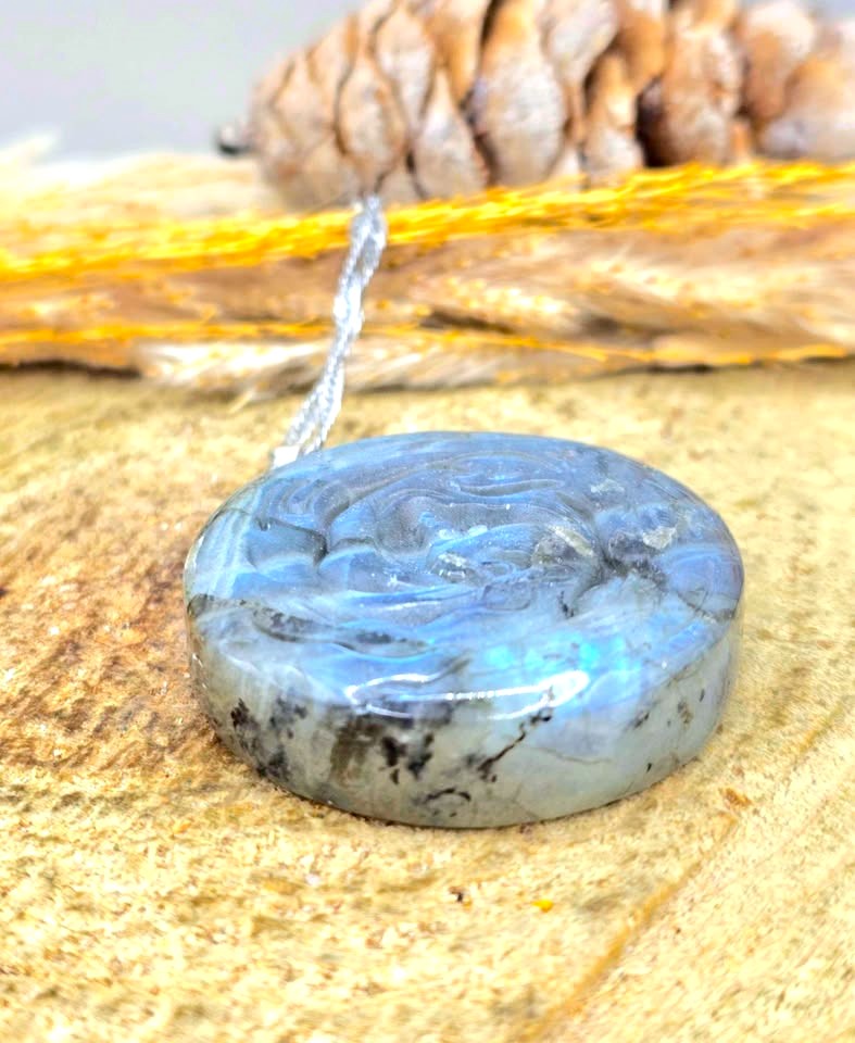 A polished round labradorite pendant with a swirling blue and gray marbled pattern, hanging from a silver cord. The pendant is displayed on a textured tan surface with a pinecone and yellow string in the blurred background.