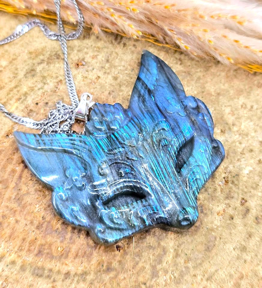 A carved blue and teal resin or stone fox head pendant necklace with a silver chain, displayed on a textured tan surface with dried grass in the background.