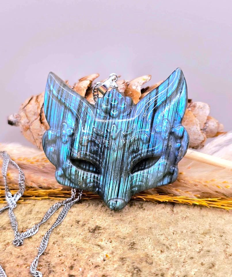 A carved fantasy fox or wolf head pendant with a metallic blue and green patina, resting on dried grass and bones with a silver chain draped in the foreground.