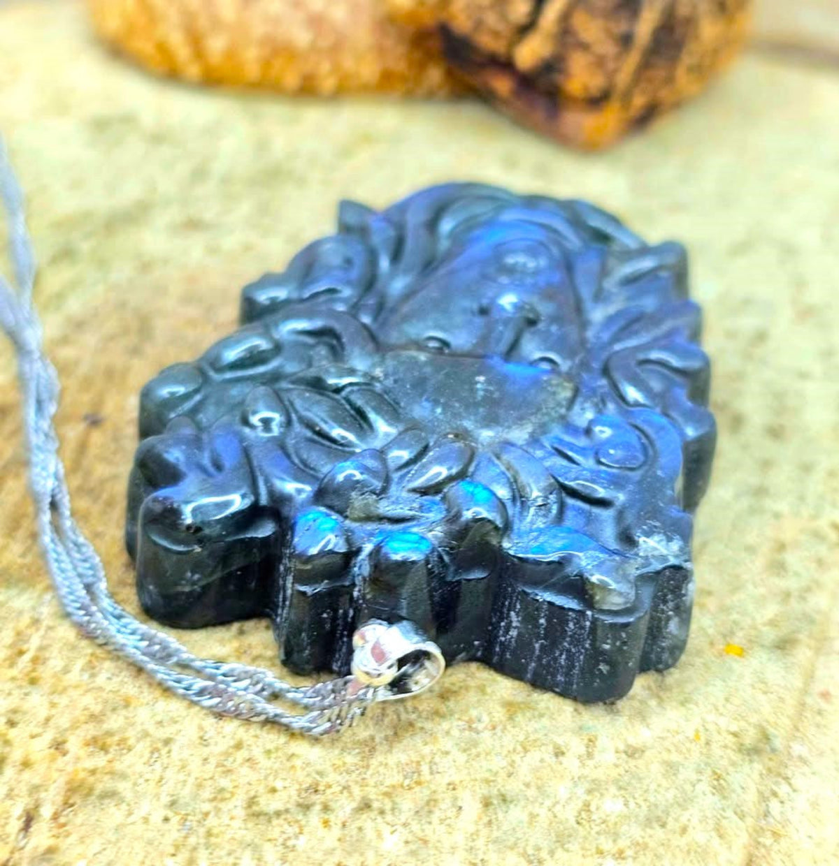 A carved dark blue labradorite pendant with a silver-toned metal cap and a gray cord, resting on a textured beige stone surface.