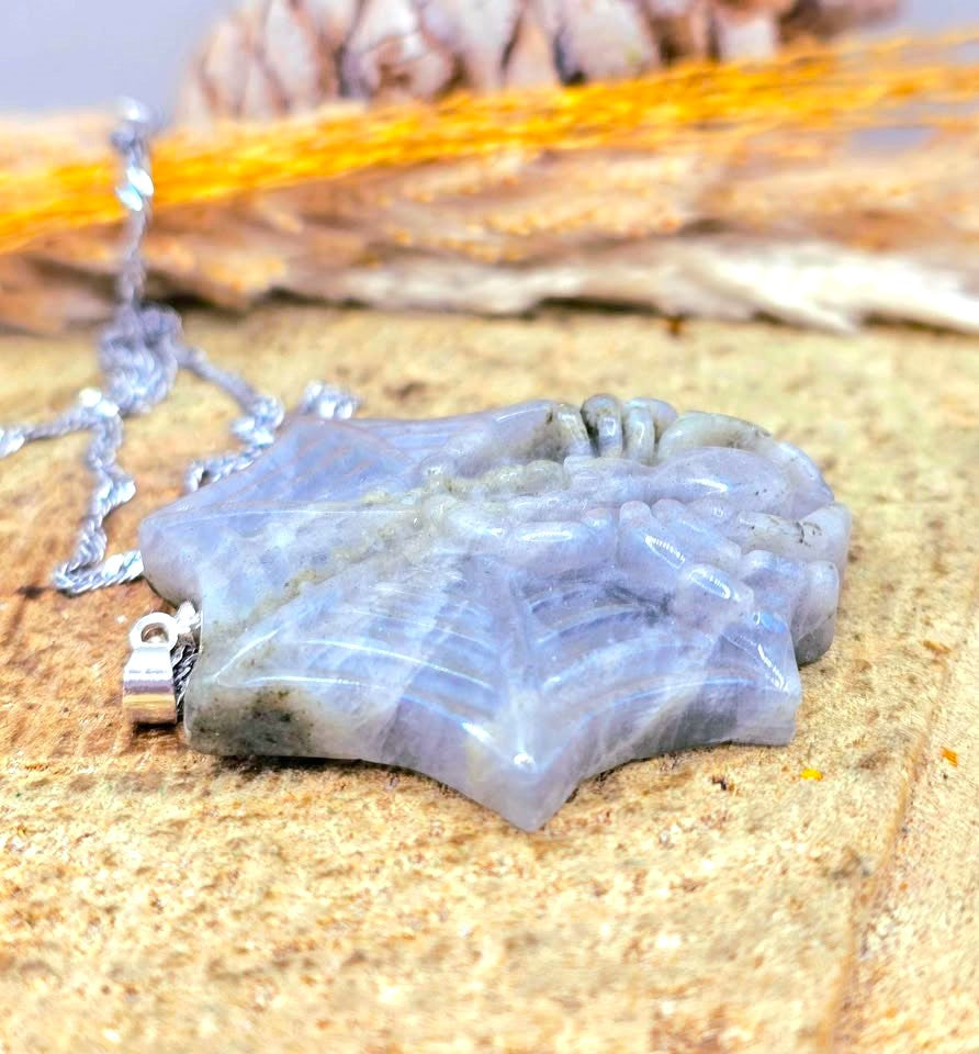 A carved stone pendant necklace with a silver bail and chain, featuring a textured, mountain-like relief design in a pale lavender-gray hue, displayed on a sandy surface.