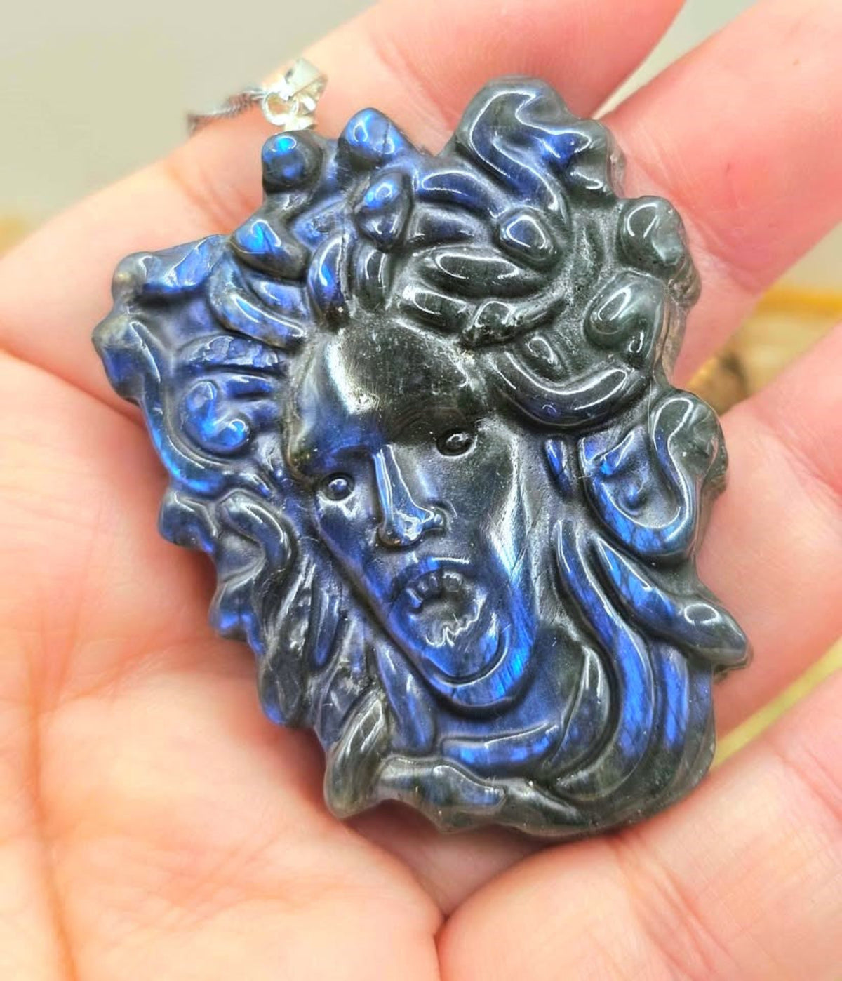 A carved labradorite pendant shaped like Medusa's face, held in a person's hand. The pendant features a dark blue and black labradorite finish with a silver bail at the top.