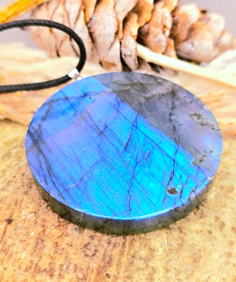 A round labradorite pendant necklace with a vibrant blue and purple iridescent sheen, resting on a rustic wooden surface with a pinecone and dried leaves in the background.