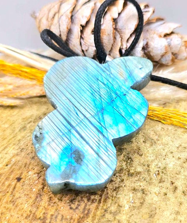 A polished labradorite crystal pendant shaped like a stylized figure, featuring a vibrant blue and green iridescent sheen, suspended from a black cord. The pendant is displayed on a textured wooden surface with a pine cone and yellow string in the background.