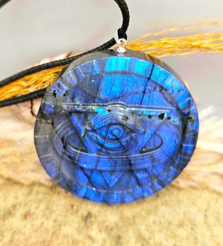 A round blue labradorite pendant necklace featuring an engraved eye of providence symbol, suspended on a black cord against a neutral background with dried grass.