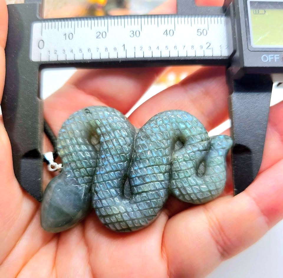 A carved snake-shaped pendant or charm with a textured scale pattern, held in a person's hand under a digital caliper to show its dimensions. The pendant has a gray-green iridescent finish.